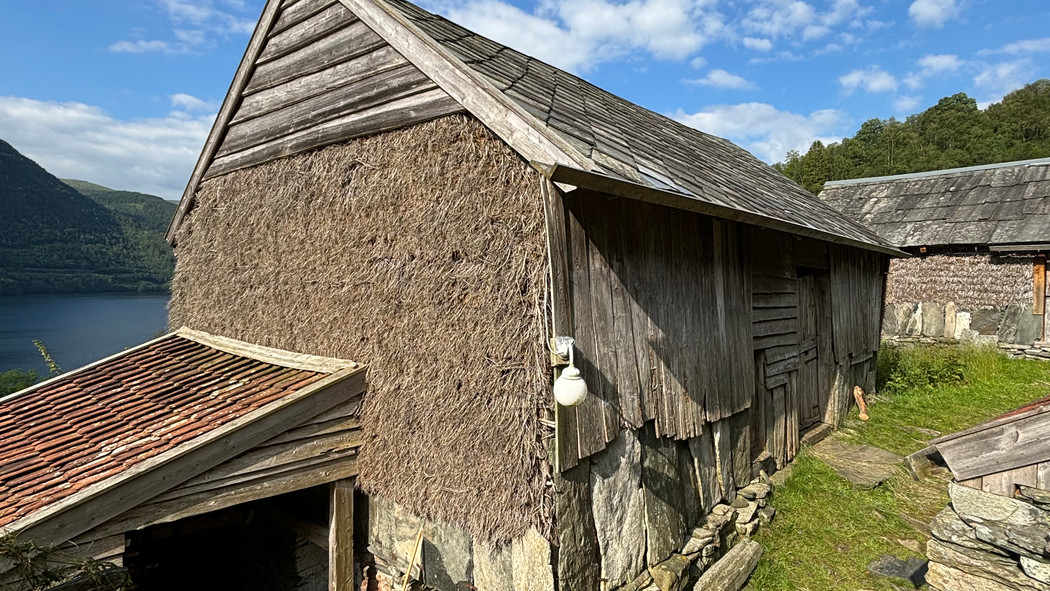 Løe fra 1878 med skifertak og kledning av tre og halm. Beliggende på gårdstun med fjordlandskap og himmel i bakgrunnen.